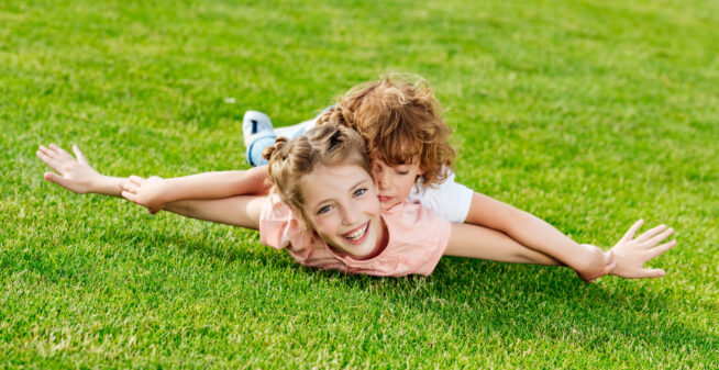 adorable brother and sister with arms outstretched lying on green grass in park