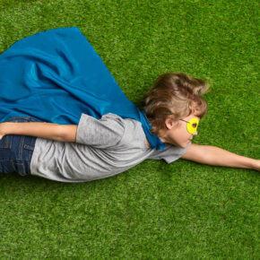 Little superhero flying over grass Little boy in cape and mask lying in flying pose on green lawn while pretending to be superhero
