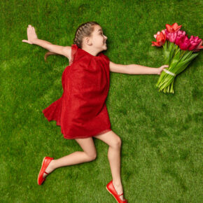 Playful girl with bouquet on green meadow From above of charming girl in red dress lying on green lawn with bouquet of tulips